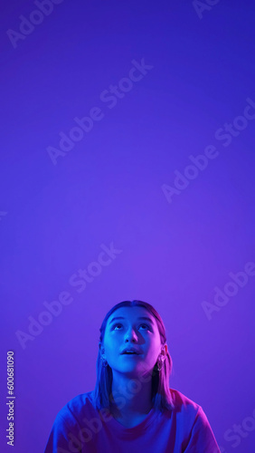 Color light portrait. Amazed girl. Neon surprised woman looking up with interest at advertisement area isolated on blue pink empty space background.