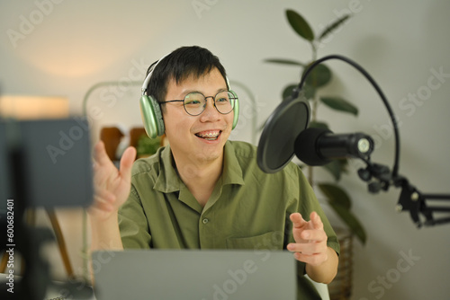 Asian man radio host using laptop and talking through microphone to recording podcast in small home studio