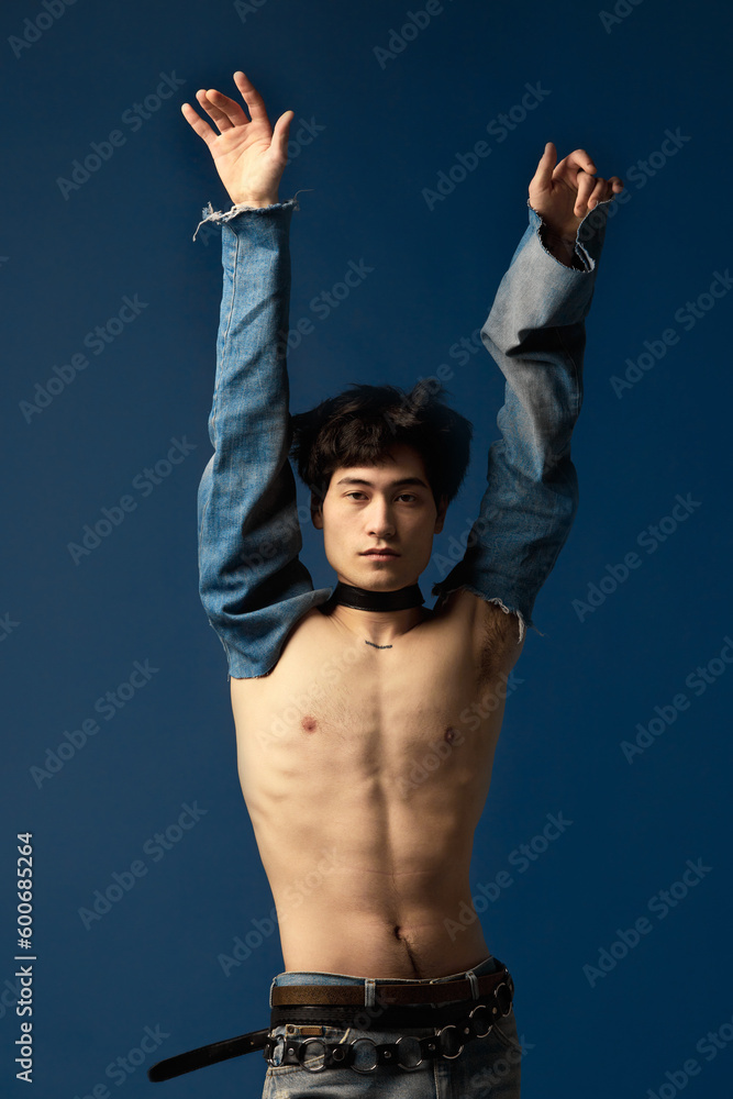 Portrait of young man posing shirtless with jeans sleeve elements against blue studio background. Extravagant design. Concept of men's fashion, extraordinary style, art, beauty, trends