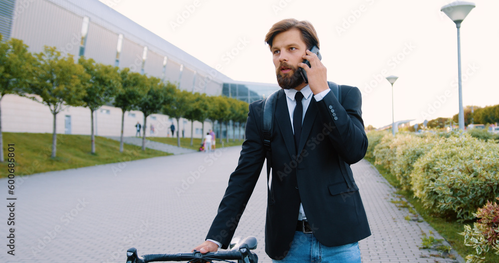 Attractive confident purposeful 30-aged bearded stylish man in black jacket and jeans with backpack walking on pedestrian path with his bicycle and talking on smartphone on modern impressive building Attractive confident purposeful 30-aged bearded stylish man in black jacket and jeans with backpack walking on pedestrian path with his bicycle and talking on smartphone on modern impressive building