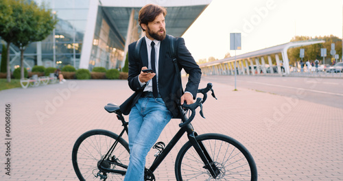 Handsome confident positive young bearded man standing near his bike using phone on the city airport background