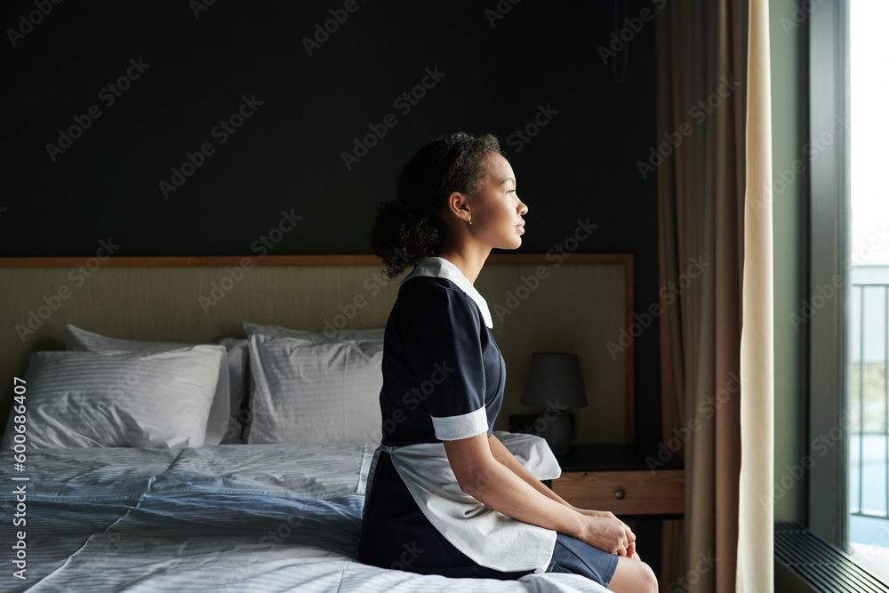 Young tired chambermaid in uniform sitting on double bed with two pillows in hotel room and looking through large window in front of her Young tired chambermaid in uniform sitting on double bed with two pillows in hotel room and looking through large window in front of her