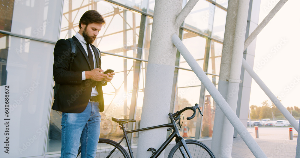 Handsome confident purposeful young bearded man standing near glass wall with his bike and using phone to write message ,front view