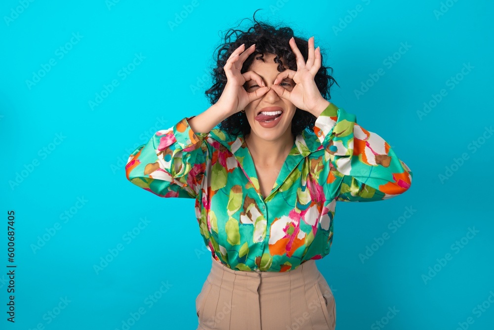 Young arab woman wearing colorful shirt over blue background doing ok gesture like binoculars sticking tongue out, eyes looking through fingers. Crazy expression. Young arab woman wearing colorful shirt over blue background doing ok gesture like binoculars sticking tongue out, eyes looking through fingers. Crazy expression.