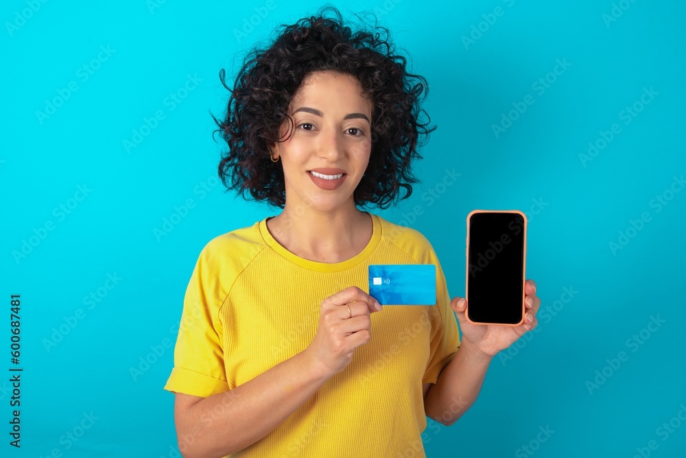 Photo of adorable Young arab woman wearing yello T-shirt over blue background holding credit card and Smartphone. Reserved for online purchases Photo of adorable Young arab woman wearing yello T-shirt over blue background holding credit card and Smartphone. Reserved for online purchases