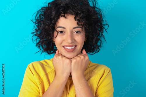 Satisfied Young arab woman wearing yello T-shirt over blue background touches chin with both hands, smiles pleasantly, rejoices good day with lover