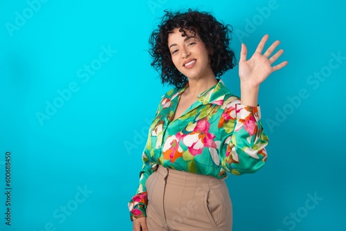 young arab woman wearing colorful shirt over blue background Waiving saying hello happy and smiling, friendly welcome gesture.