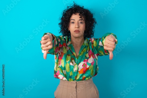 Fototapeta young arab woman wearing colorful shirt over blue background being upset showing thumb down with two hands