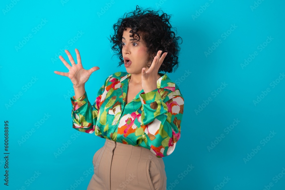 young arab woman wearing colorful shirt over blue background shouts loud, keeps eyes opened and hands tense. young arab woman wearing colorful shirt over blue background shouts loud, keeps eyes opened and hands tense.