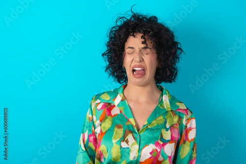 young arab woman wearing colorful shirt over blue background sticking tongue out happy with funny expression. Emotion concept.