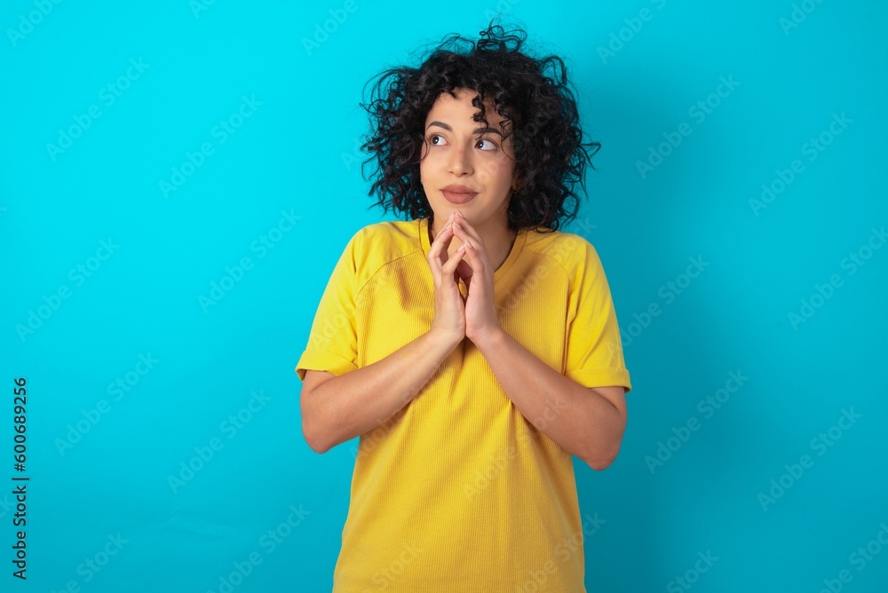 young arab woman wearing yellow T-shirt over blue background steepled fingers and looks mysterious aside has great evil plan in mind young arab woman wearing yellow T-shirt over blue background steepled fingers and looks mysterious aside has great evil plan in mind