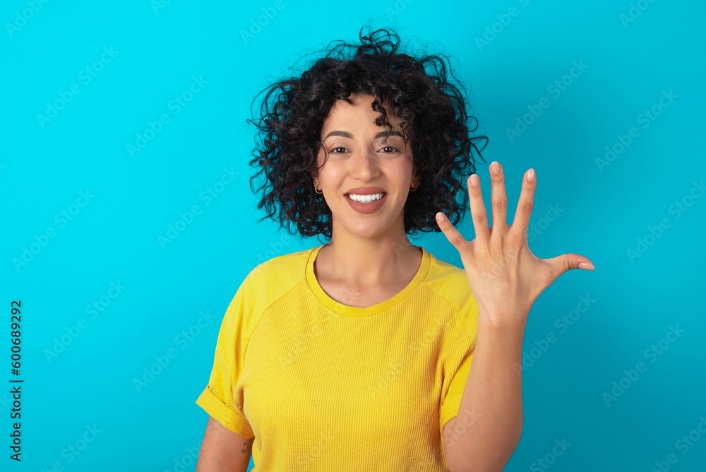 young arab woman wearing yellow T-shirt over blue background smiling and looking friendly, showing number five or fifth with hand forward, counting down young arab woman wearing yellow T-shirt over blue background smiling and looking friendly, showing number five or fifth with hand forward, counting down