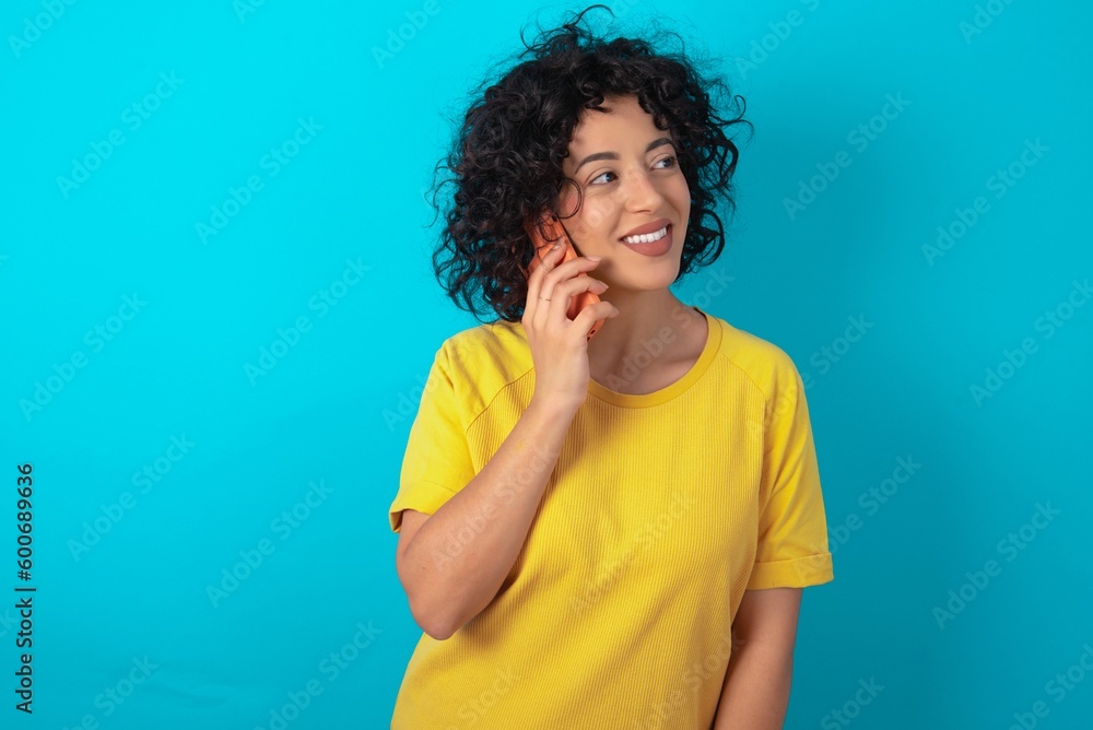 Pleasant looking happy young arab woman wearing yellow T-shirt over blue background has nice telephone conversation and looks aside, has nice mood and smiles positively while talks via cell phone Pleasant looking happy young arab woman wearing yellow T-shirt over blue background has nice telephone conversation and looks aside, has nice mood and smiles positively while talks via cell phone