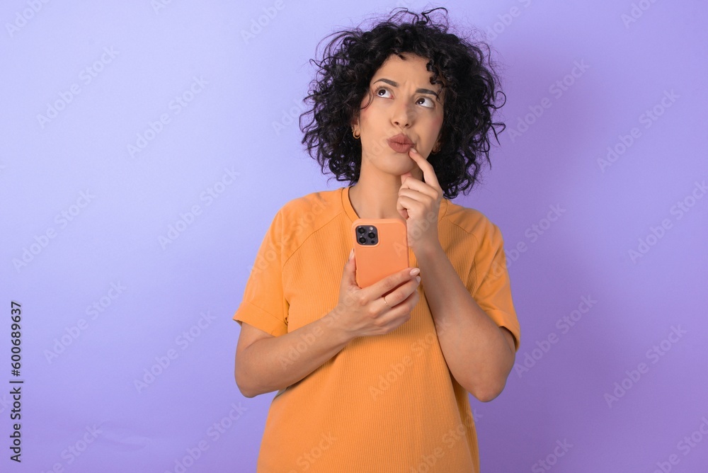 young arab woman wearing yellow T-shirt over purple background thinks deeply about something, uses modern mobile phone, tries to made up good message, keeps index finger near lips. young arab woman wearing yellow T-shirt over purple background thinks deeply about something, uses modern mobile phone, tries to made up good message, keeps index finger near lips.