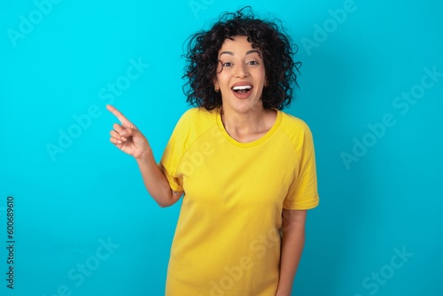 young arab woman wearing yellow T-shirt over blue background laughs happily points away on blank space demonstrates shopping discount offer, excited by good news or unexpected sale. © Jihan