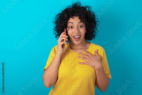 Smiling young arab woman wearing yellow T-shirt over blue background talks via cellphone, enjoys pleasant great conversation. People, technology, communication concept
