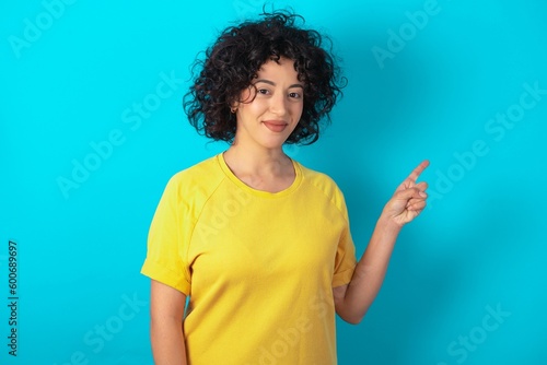 young arab woman wearing yellow T-shirt over blue background points to side on blank space demonstrates advertisement. People and promotion concept