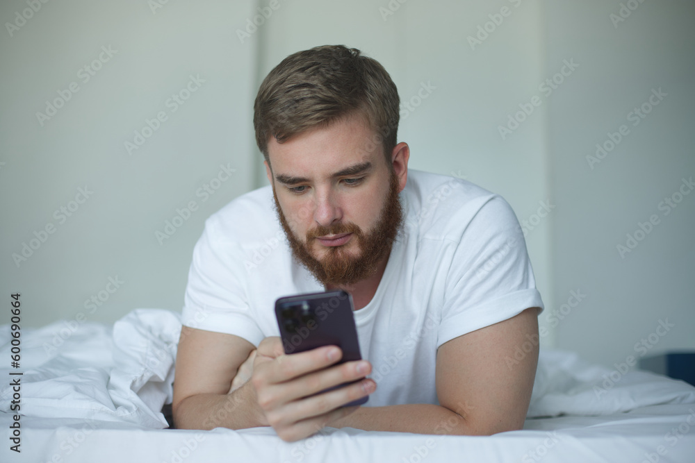 Young bearded man is using cell mobile phone at home in bed in bedroom, looking at screen of smartphone Young bearded man is using cell mobile phone at home in bed in bedroom, looking at screen of smartphone