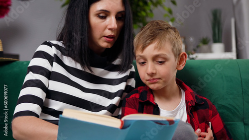 good-looking stylish modern contented dark-haired mom and her handsome 8-aged son which spending their free time at reading interesting book story in contemporary living-room photo