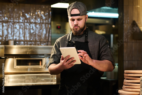 Fototapeta Male cook looking through online recipes on tablet screen while standing by work