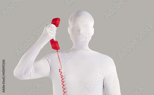 Person in faceless bodysuit disguise makes anonymous incognito phone call from unknown number. Strange unrecognizable young man wearing white spandex costume holding red vintage telephone receiver photo