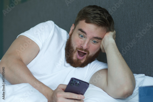 Young bearded man is using cell mobile phone at home in bed in bedroom, looking at screen of smartphone 