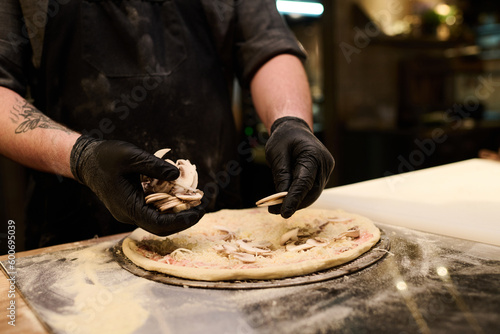 Close-up of young chef putting fresh chopped mushrooms on flatbread for Italian pizza while standing by table in the kitchen and cooking