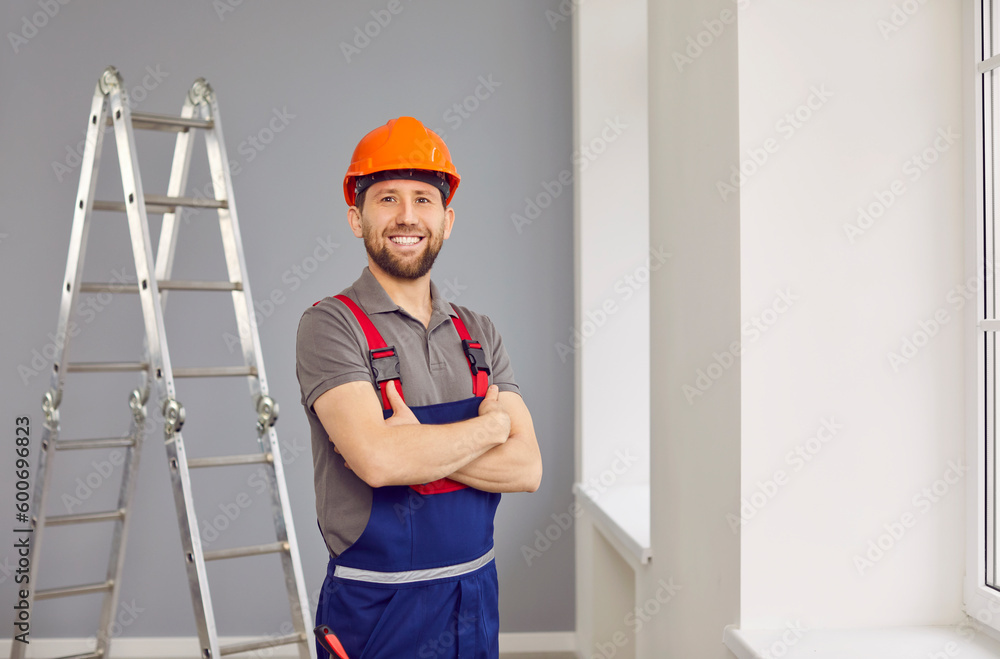 Portrait of a construction worker or repairman in the house. Happy repair man wearing a safety helmet and workwear overalls standing with folded arms in the room, with a stepladder in the background Portrait of a construction worker or repairman in the house. Happy repair man wearing a safety helmet and workwear overalls standing with folded arms in the room, with a stepladder in the background