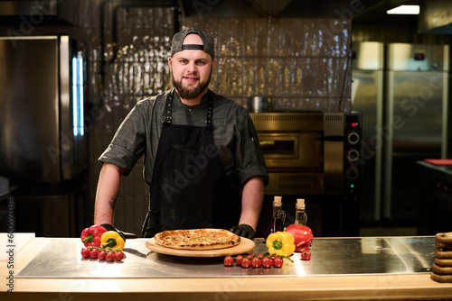 Happy young successful chef and owner of restaurant or pizzeria looking at camera while standing by table with appetizing pizza in the kitchen