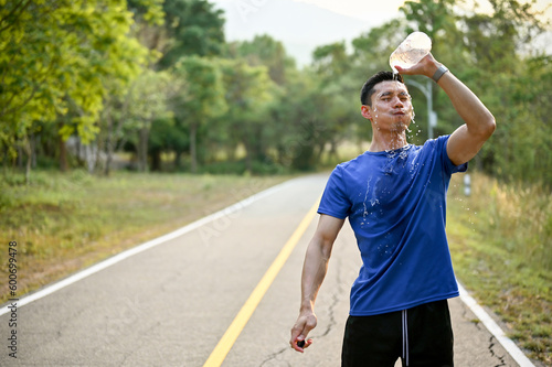 Tired Asian male runner pouring water from a bottle on his face, refreshing himself with water