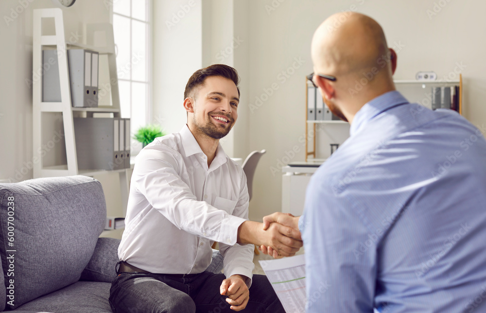 Obraz premium Two happy smiling men shaking hands sitting in the office celebrating success, making a good deal, business achievement, signing a contract or greeting new employee on their workplace.