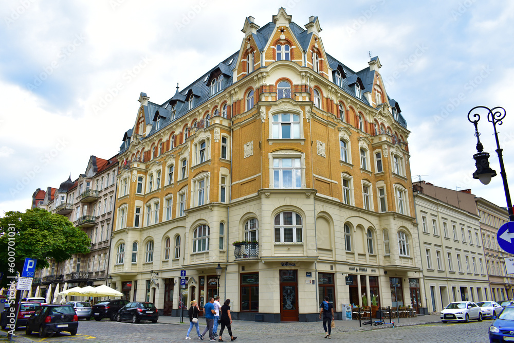 Old architectural corner building with beige and orange walls, windows and a blue roof. City street with cars and people. Summer cloudy day.  Poland, Poznan, June 2022