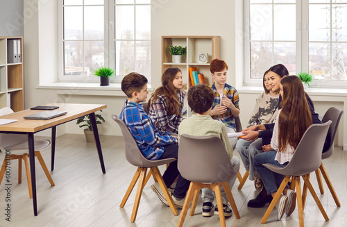 Group of elementary school children and their teacher discussing something while sitting in a circle on comfortable chairs in a modern classroom all together photo