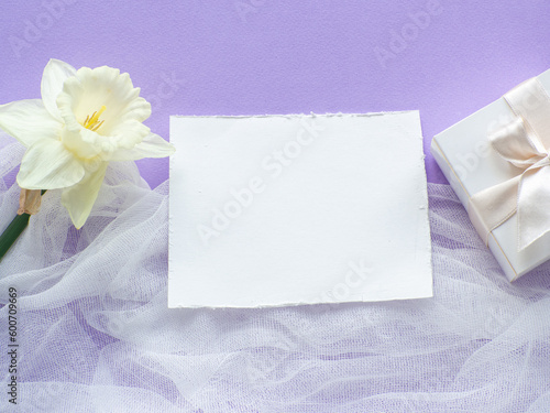Daffodils flower and gift box on lilac background with copy space. Women and Mothers Day greeting card. Place for text and narcissus on purple background. Birthday congratulations note flat lay.
