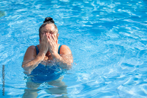 Portrait of wonderful buxom middle-aged woman wearing blue swimsuit, standing in swimming pool, wiping face with hands.
