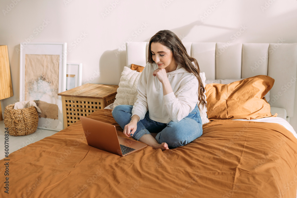 young curly girl in home casual clothes studying online at home sitting on the bed in her bedroom young curly girl in home casual clothes studying online at home sitting on the bed in her bedroom