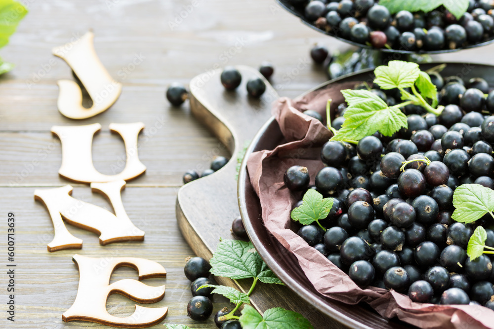 Freshly gathered big juicy black currants on brown plate closeup, black currant harvest, word July of wooden letters on table, berries outdoors, in garden Freshly gathered big juicy black currants on brown plate closeup, black currant harvest, word July of wooden letters on table, berries outdoors, in garden
