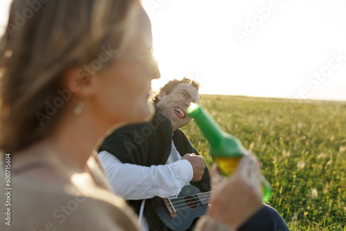 Summer time. Beautiful young couple enjoying time in field around grass. Lifestyle, love, dating, vacation concept. Woman drinking beer, man playing ukulele