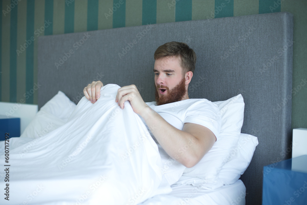 Young happy man looking at his penis under blanket, surprised guy is having good morning potency, erection in bed in bedroom  - obrazy, fototapety, plakaty 