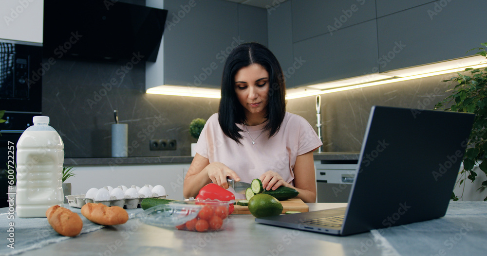 Pretty happy smiling young brunette cutting cucumber on cutting board using sharp knife and watching online culinary show on computer in modern cuisine