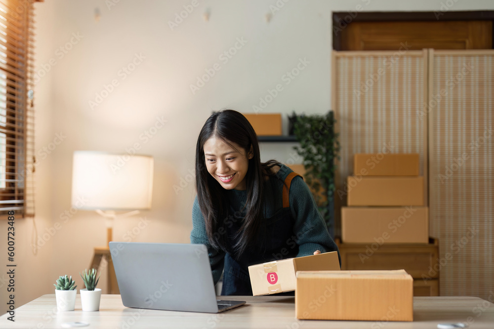 Asian businesswoman using laptop to start online delivery freelancer or salesman checking production order with laptop. SME entrepreneur working with parcel box at home Asian businesswoman using laptop to start online delivery freelancer or salesman checking production order with laptop. SME entrepreneur working with parcel box at home