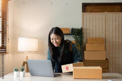 Asian businesswoman using laptop to start online delivery freelancer or salesman checking production order with laptop. SME entrepreneur working with parcel box at home