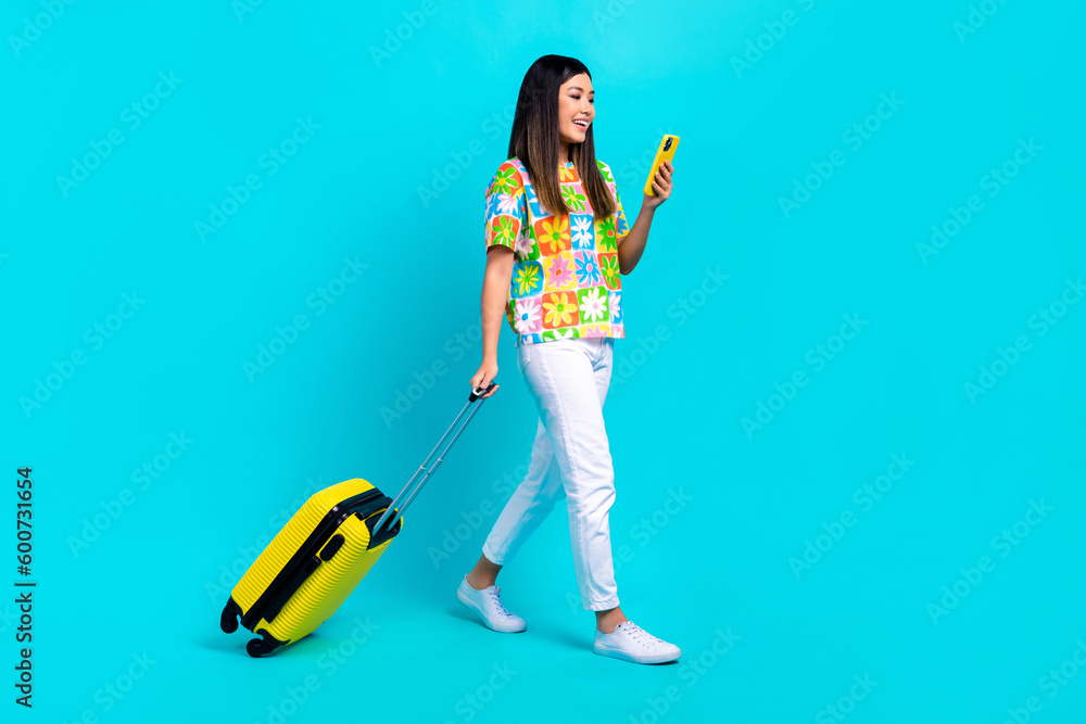 Full body cadre of young hipster chinese girl hippie hold phone walking with baggage exploring airport isolated on cyan color background Full body cadre of young hipster chinese girl hippie hold phone walking with baggage exploring airport isolated on cyan color background