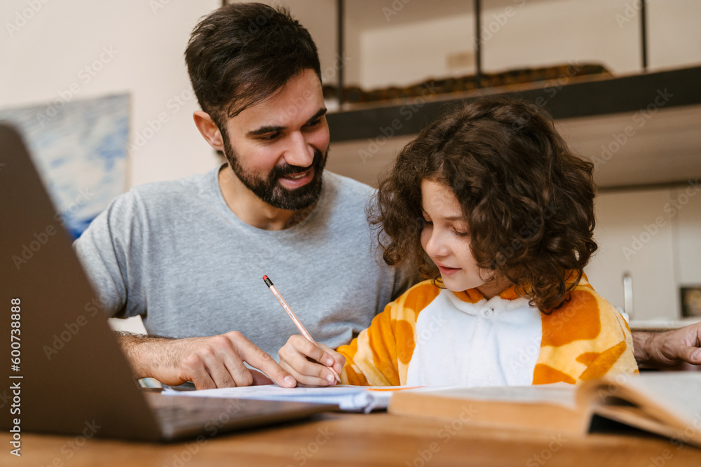 Father helping his son with homework while sitting with laptop at table Father helping his son with homework while sitting with laptop at table