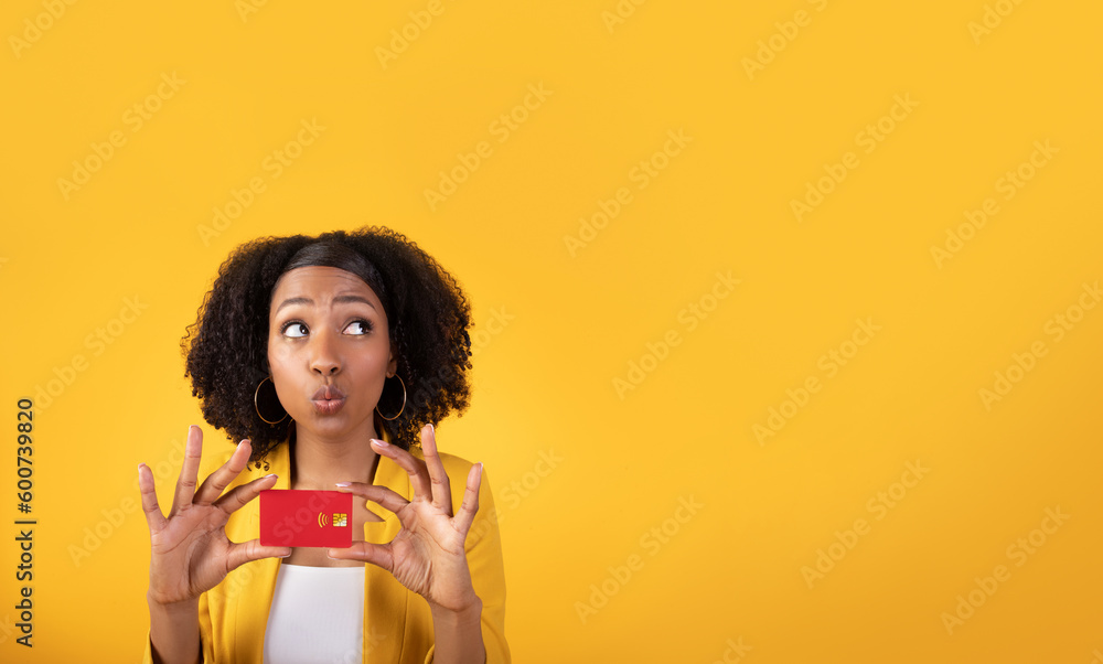Dreamy black lady with credit card enjoying online shopping, looking at free space isolated on yellow studio background Dreamy black lady with credit card enjoying online shopping, looking at free space isolated on yellow studio background