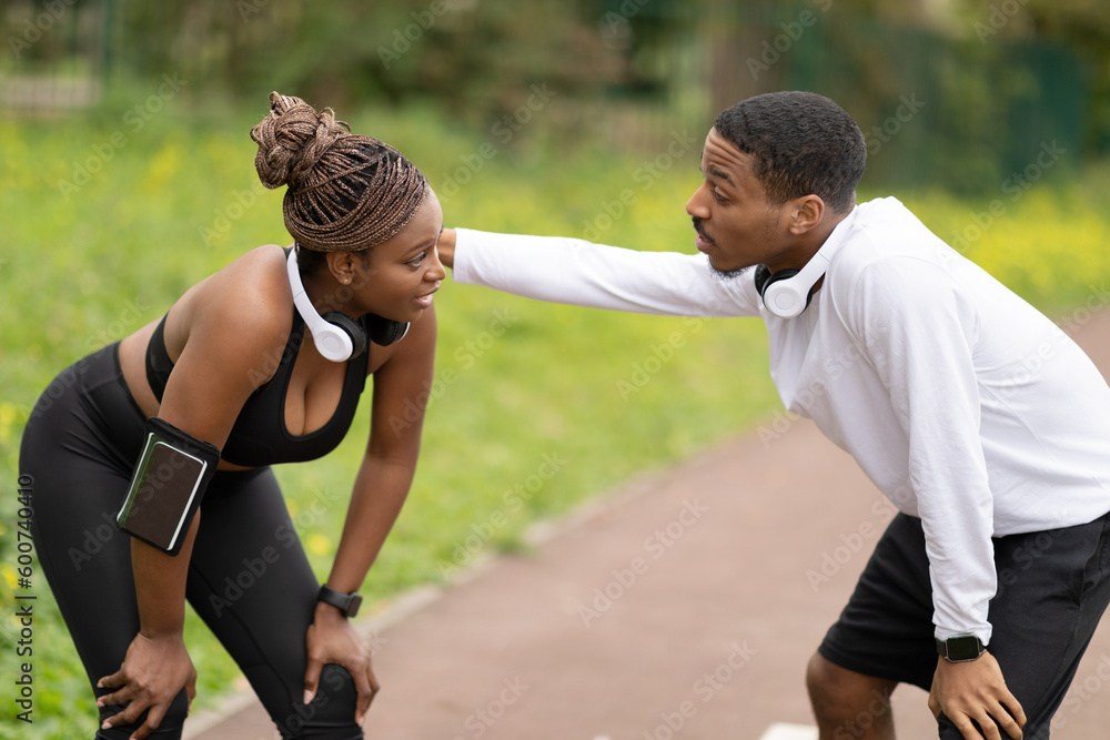 Tired cheerful young african american couple in sportswear having rest after jogging in park