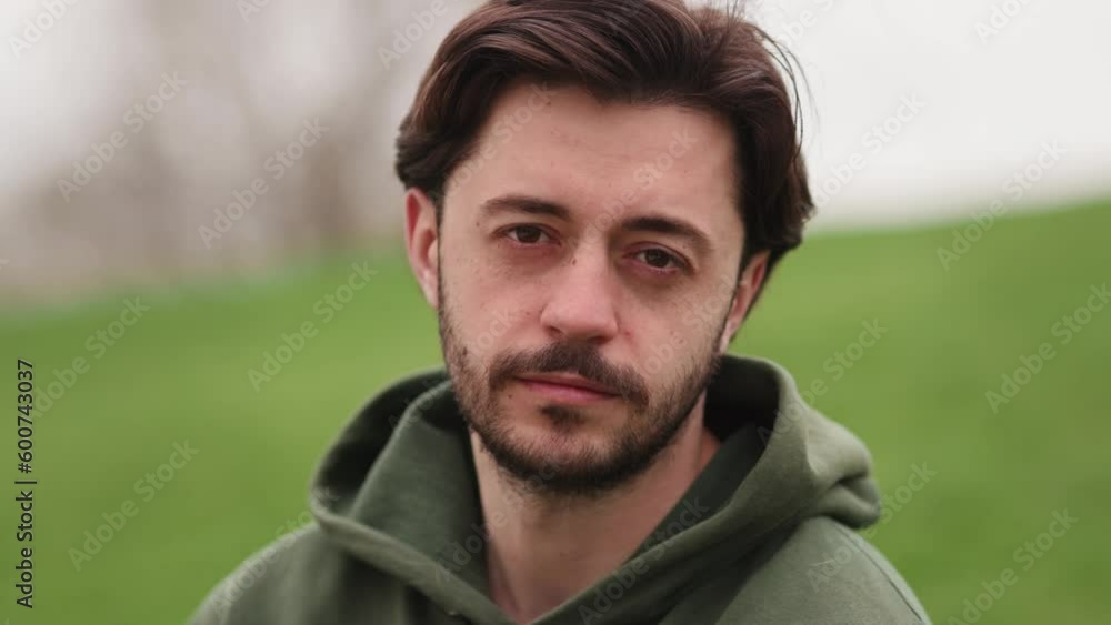 Serious young man sitting on the grass in the park turns his head and looks at the camera. A brunette man in a green sweater is relaxing outdoors in the park. Serious young man sitting on the grass in the park turns his head and looks at the camera. A brunette man in a green sweater is relaxing outdoors in the park.