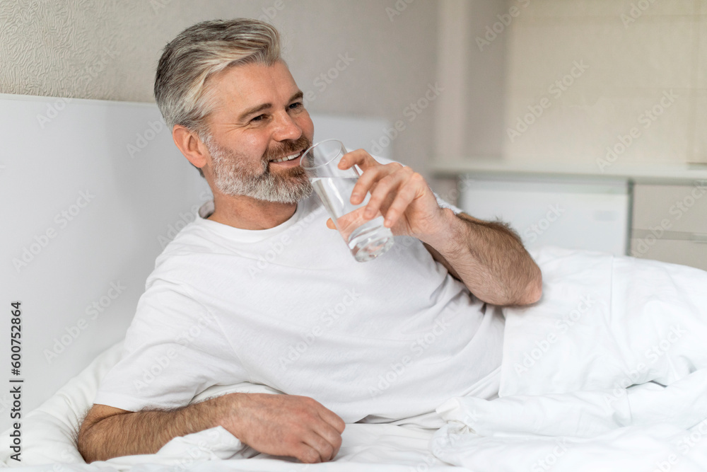 Positive european man sitting in bed, holding glass of water Positive european man sitting in bed, holding glass of water