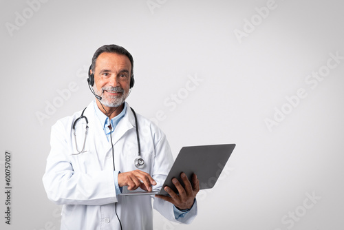Glad friendly european senior man doctor in white coat with stethoscope, headphones typing on laptop
