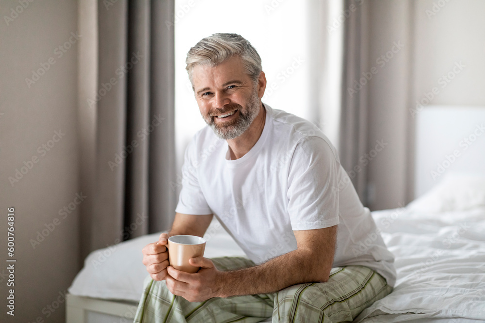 Happy middle aged man sitting on bed, drinking coffee Happy middle aged man sitting on bed, drinking coffee